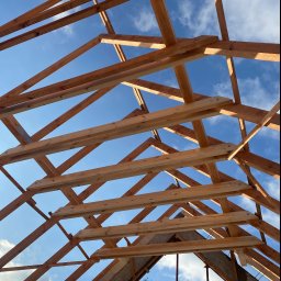 Ihor Kadynskyi - Konstrukcja drewniana dachu w trakcie budowy, widok od dołu na niebieskie niebo z chmurami, elementy połączone metalowymi płytkami i śrubami.