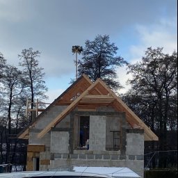 Ihor Kadynskyi - Surowy stan budowy domu z betonu komórkowego, widoczny drewniany szkielet dachu i wiecha na szczycie konstrukcji, częściowo zasłonięty przez zaparkowany samochód w zimowy dzień.