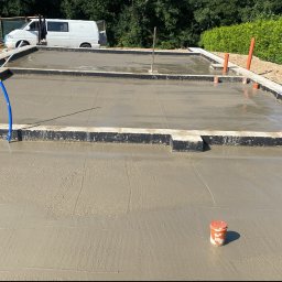 Ihor Kadynskyi - Świeżo wylana, mokra płyta fundamentowa z widocznymi kształtkami szalunkowymi, wystającymi rurami instalacyjnymi i zaparkowanym białym vanem w tle.