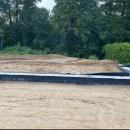 Przygotowany teren pod budowę domu z widocznymi fundamentami zabezpieczonymi czarną folią izolacyjną, dwie butelki wody stojące na fundamencie, w tle ziemia wyrównana pod wylewkę i prowizoryczna...