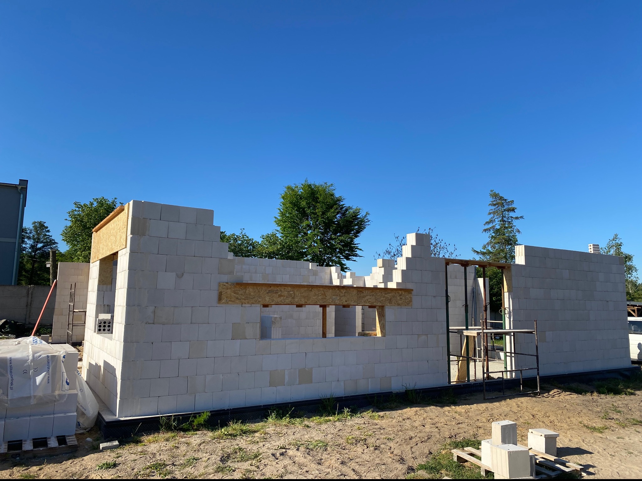 Wznoszone ściany parterowego domu z białych bloczków, widoczne otwory okienne z drewnianym szalunkiem, na ziemi paleta z zapasem bloczków, rusztowanie w tle, błękitne niebo.