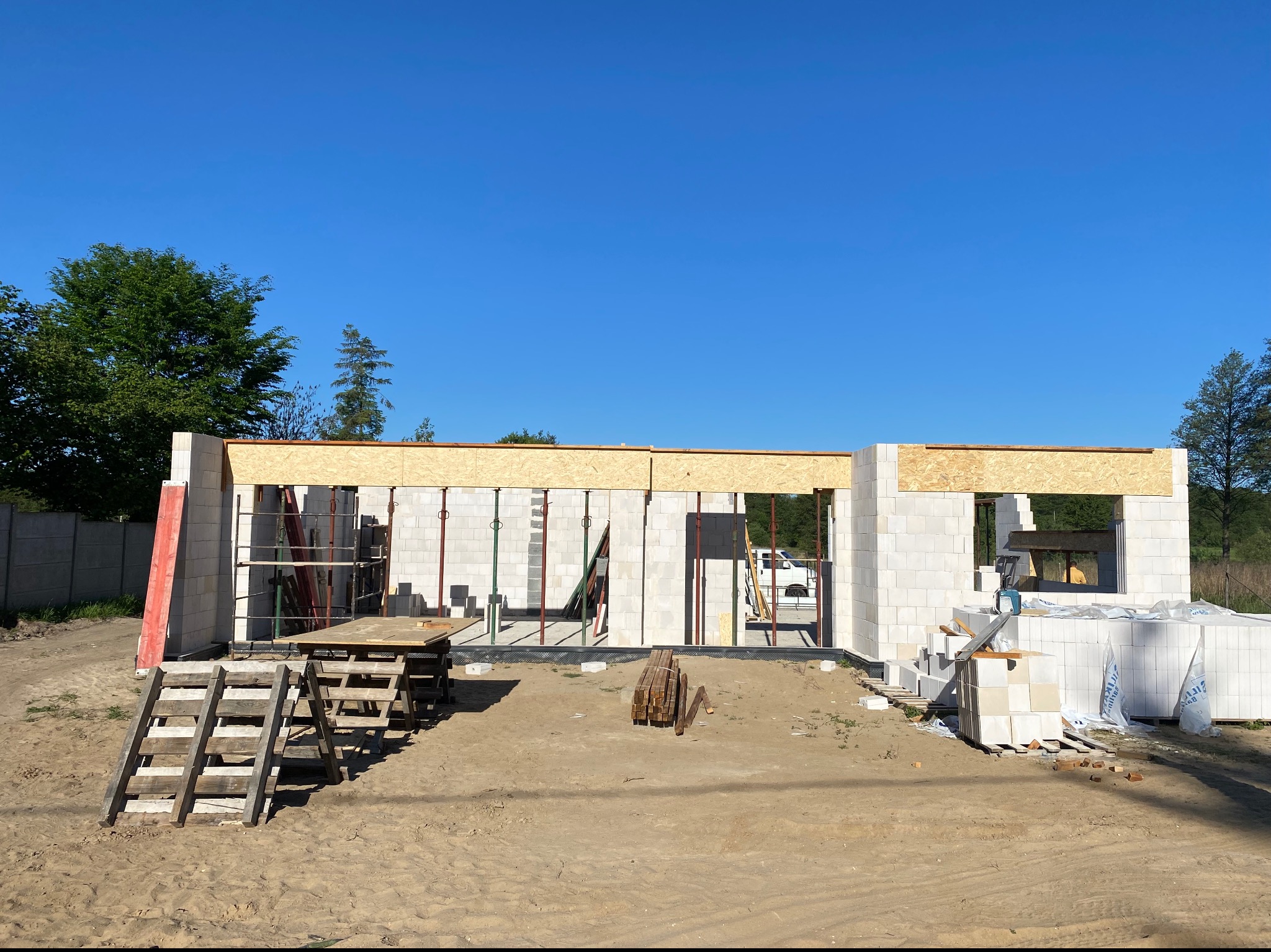 Wznoszenie ścian parterowego domu z bloczków betonu komórkowego, widoczne stemple podtrzymujące nadproża, przygotowane materiały budowlane i drewniane schodki na placu budowy w słoneczny dzień.