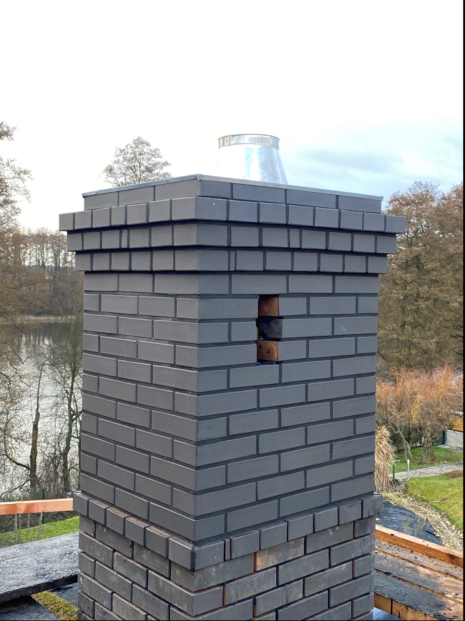 Komina z ciemnej cegły klinkierowej z metalowym wkładem, widoczny fragment uszkodzonej cegły, w tle jezioro i drzewa.