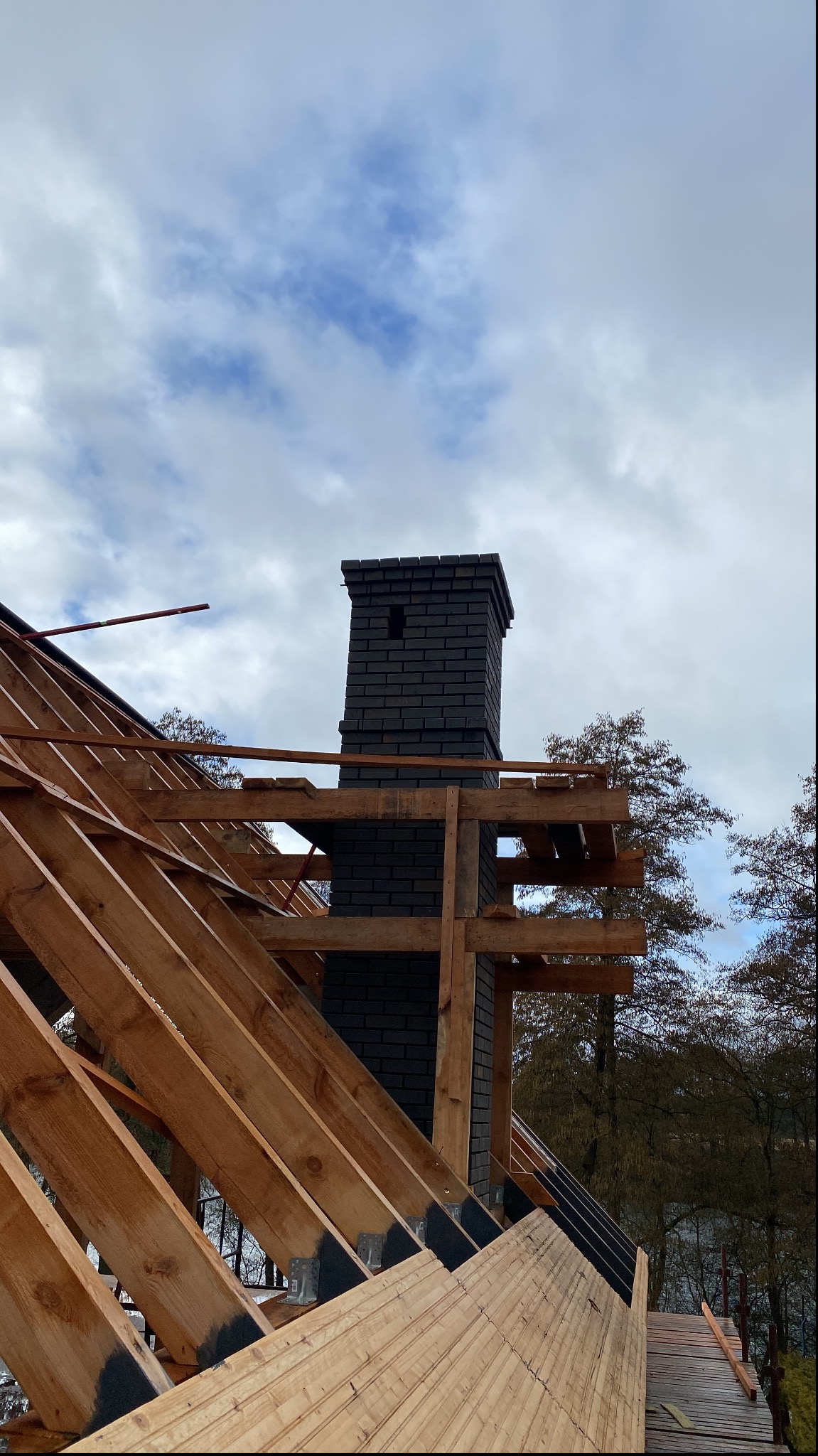 Konstrukcja dachu z drewnianymi krokwiami i kominem z ciemnej cegły, widok z dołu na tle pochmurnego nieba i drzew