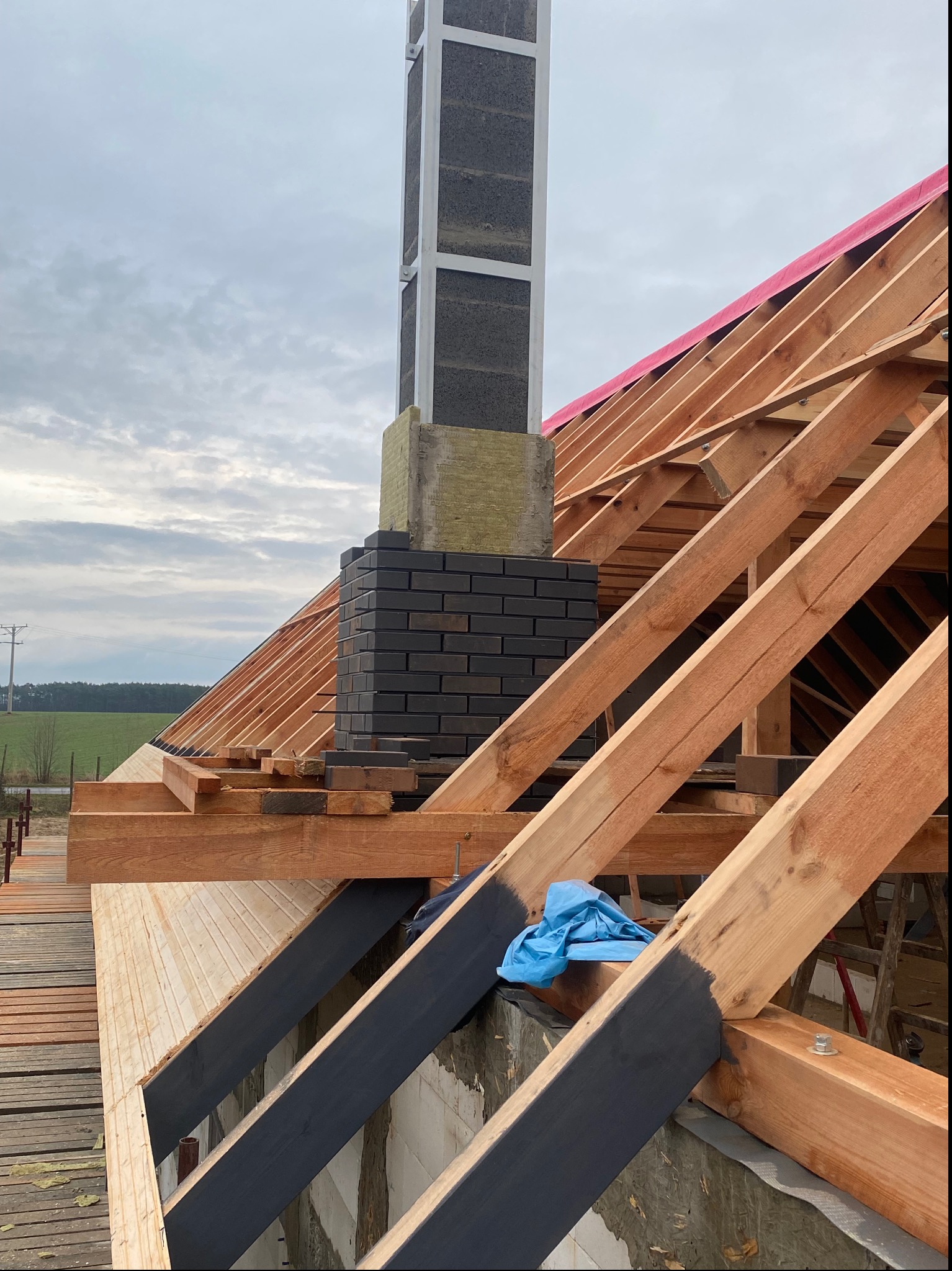 Wznoszenie komina z czarnej cegły klinkierowej na drewnianej konstrukcji dachu z widoczną membraną w kolorze różowym, w tle krajobraz wiejski pod pochmurnym niebem.