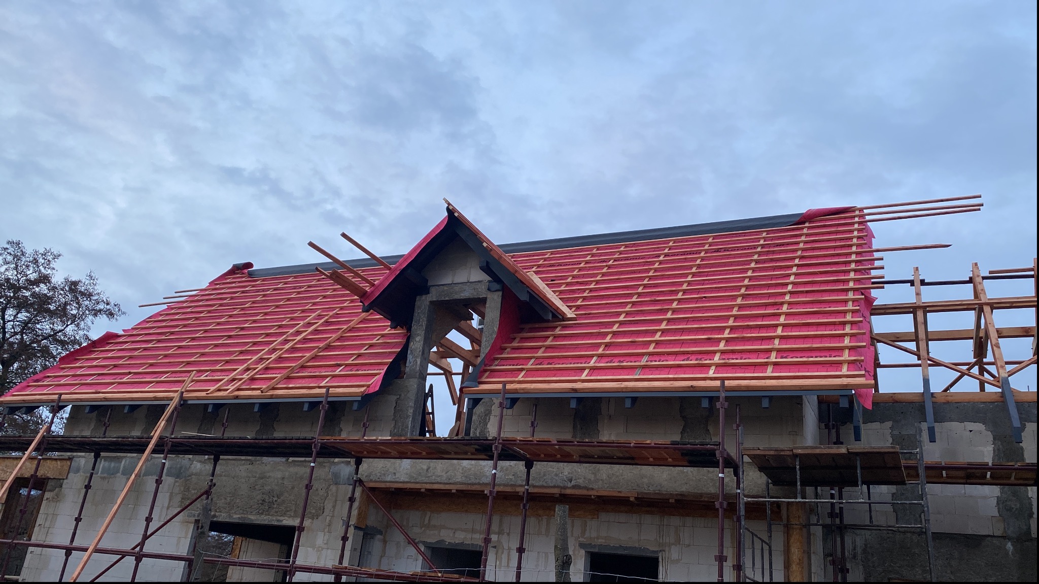 Budowa domu: widok na dach w trakcie krycia z widoczną membraną w kolorze czerwonym i drewnianą konstrukcją łat, rusztowanie w tle.