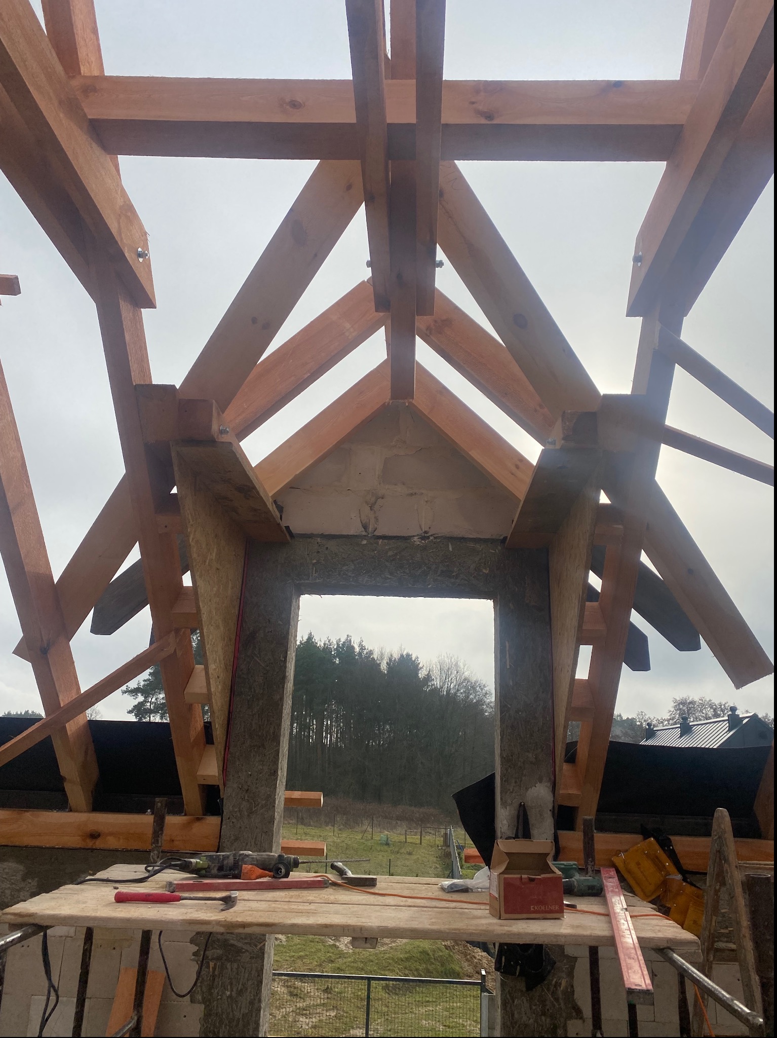 Konstrukcja drewniana dachu w trakcie budowy, widoczne krokwie i deski, narzędzia na prowizorycznym rusztowaniu, w tle las i pochmurne niebo.