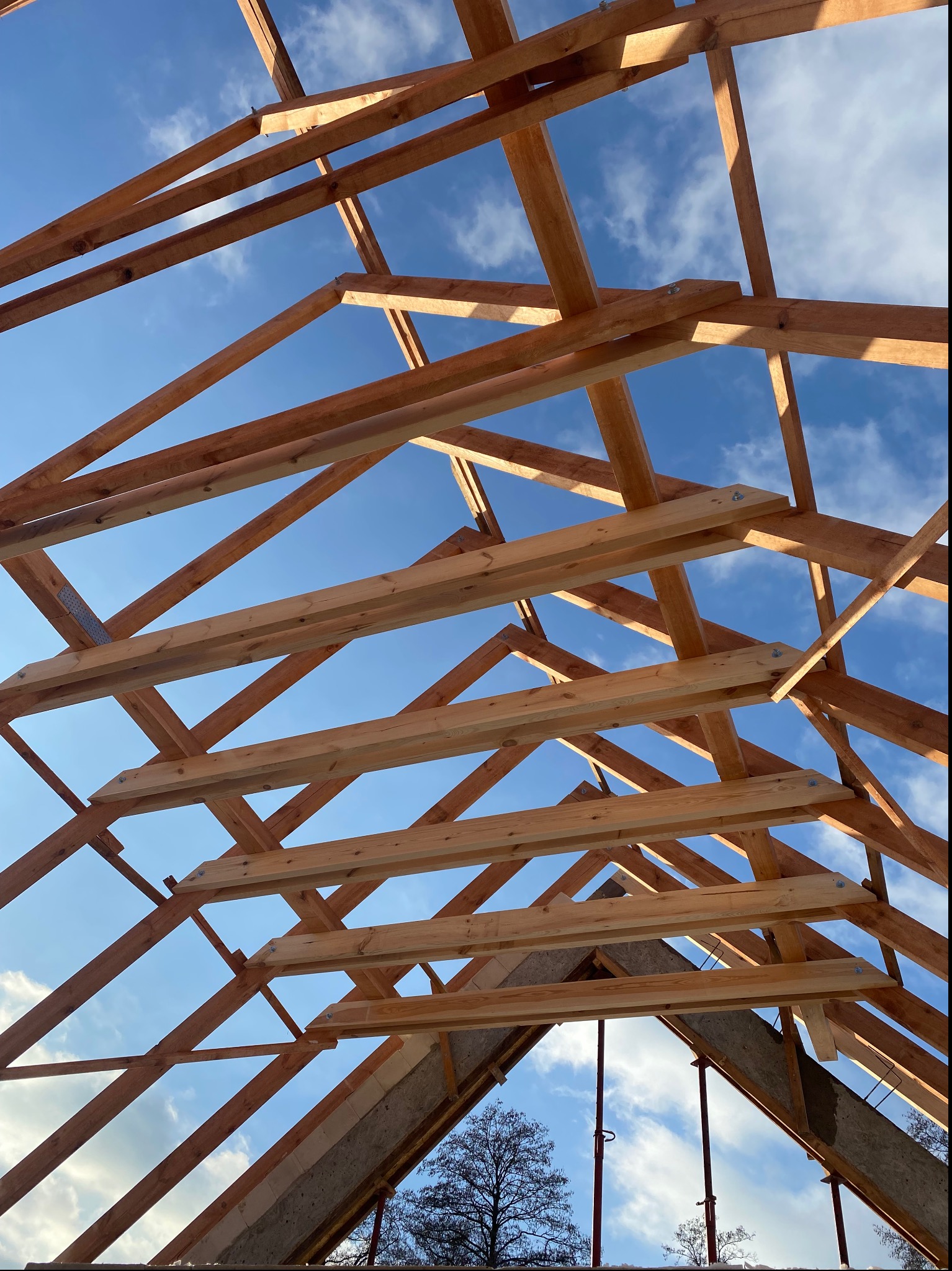 Konstrukcja drewniana dachu w trakcie budowy, widok od dołu na niebieskie niebo z chmurami, elementy połączone metalowymi płytkami i śrubami.