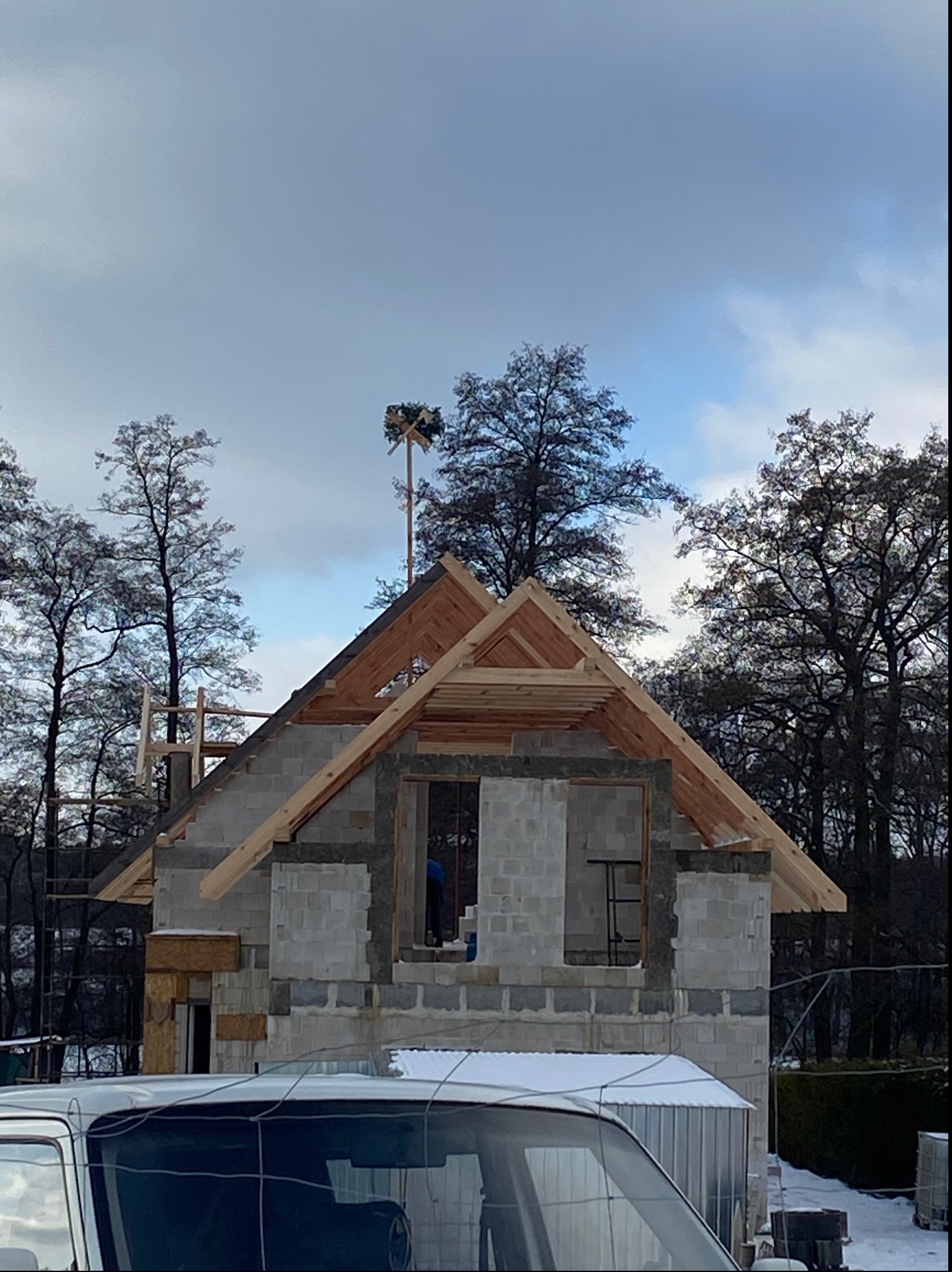 Surowy stan budowy domu z betonu komórkowego, widoczny drewniany szkielet dachu i wiecha na szczycie konstrukcji, częściowo zasłonięty przez zaparkowany samochód w zimowy dzień.