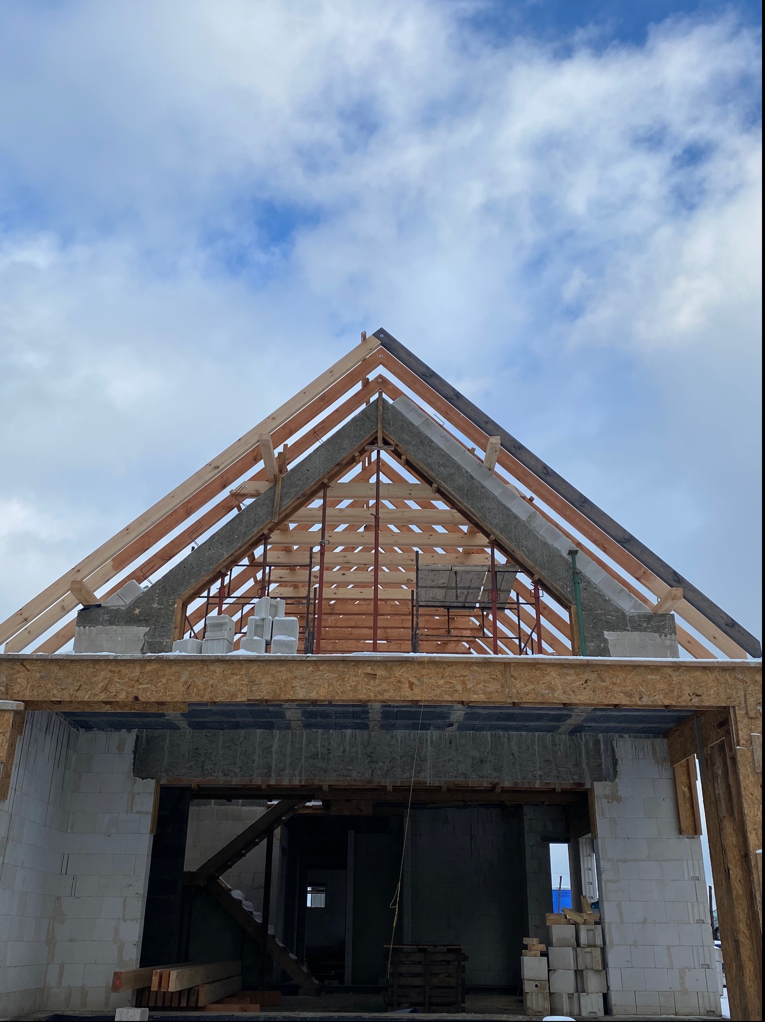 Surowy stan domu w trakcie budowy: widoczne ściany z białych bloczków, drewniana konstrukcja dachu z izolacją, w tle błękitne, pochmurne niebo.
