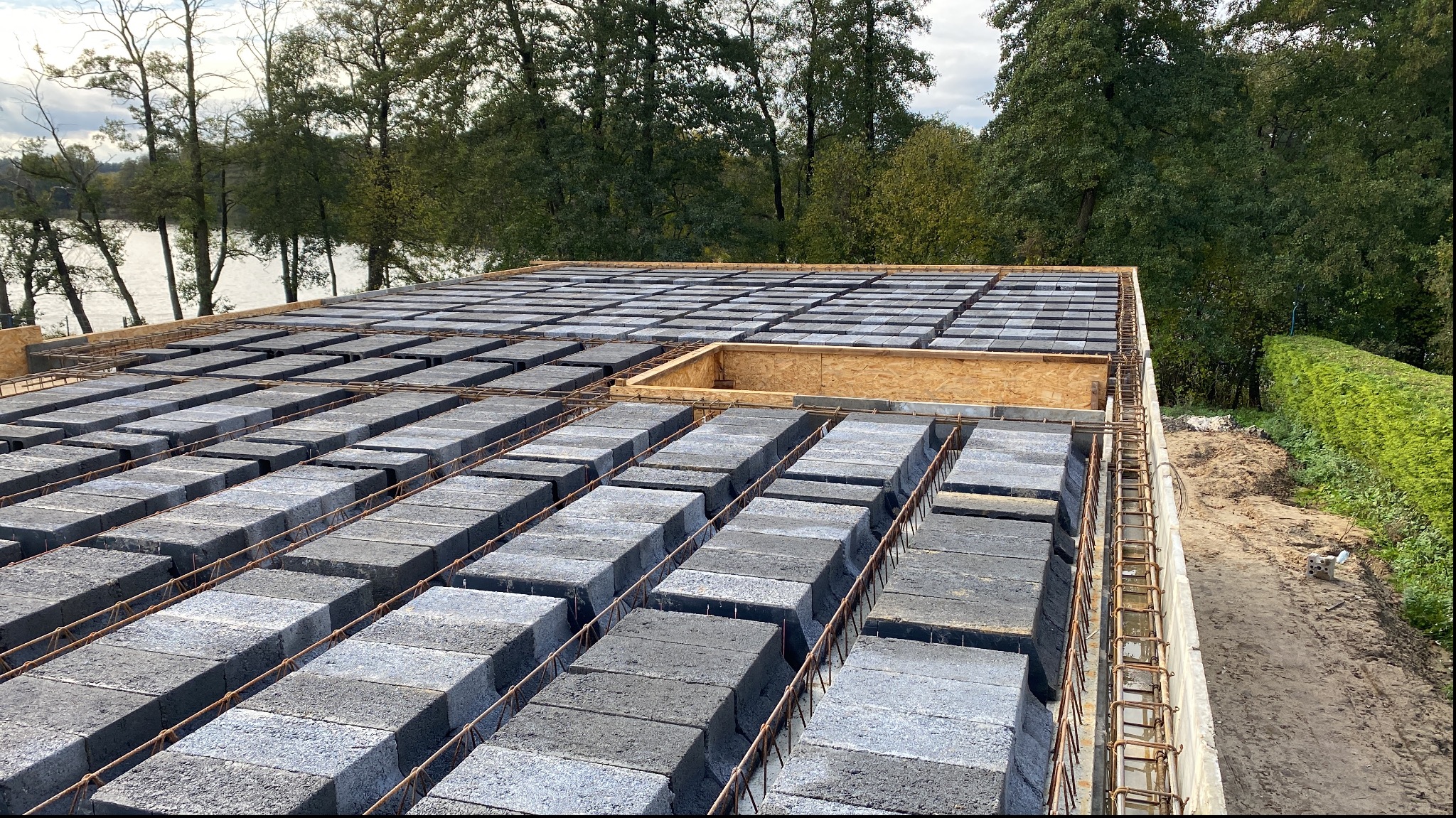 Wznoszenie stropu Teriva z pustaków szalunkowych na tle zieleni i jeziora, widoczny szalunek otworu w stropie.