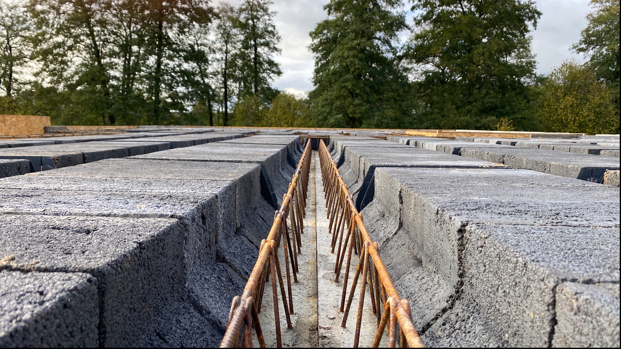 Szare bloczki fundamentowe ułożone rzędami z widocznym zbrojeniem w szczelinie, drzewa w tle.