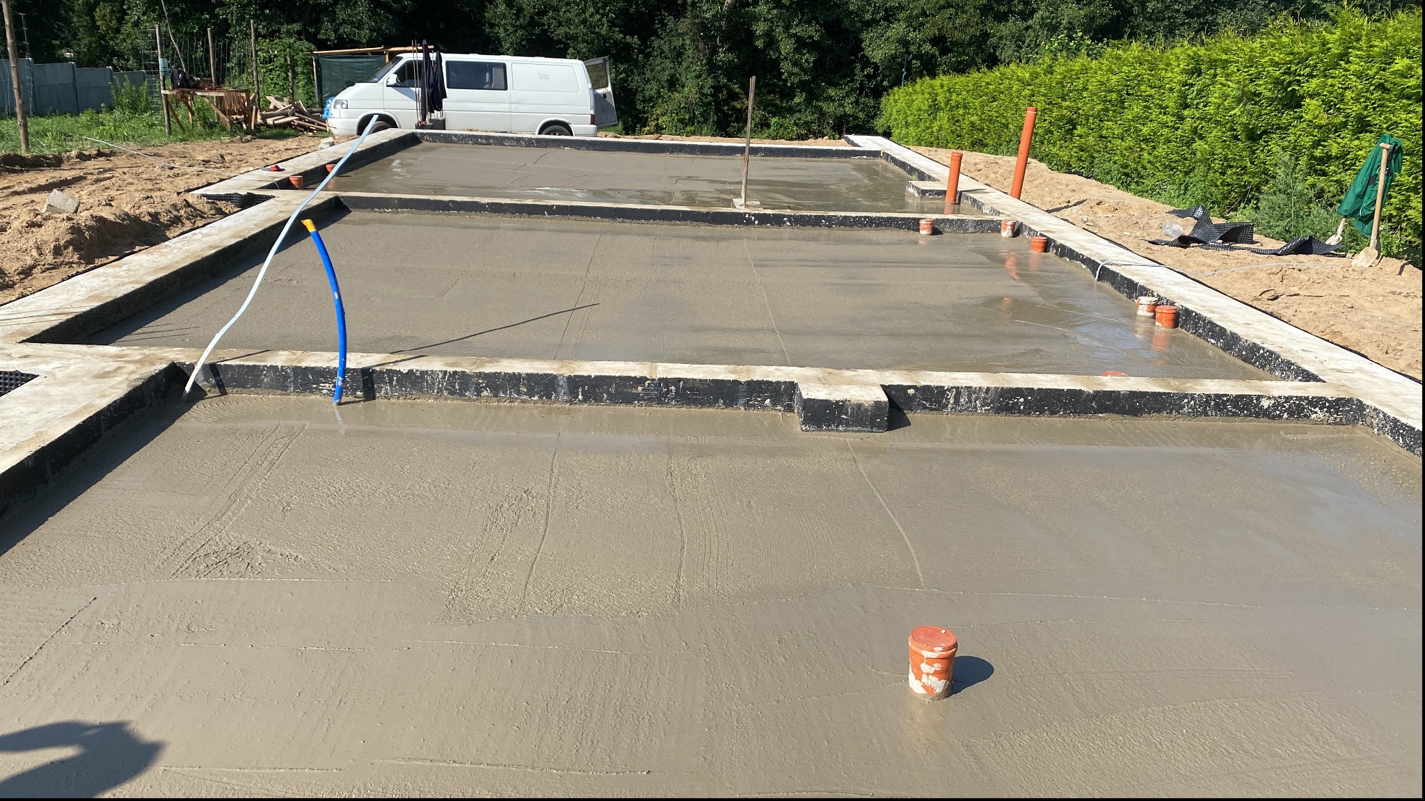 Świeżo wylana, mokra płyta fundamentowa z widocznymi kształtkami szalunkowymi, wystającymi rurami instalacyjnymi i zaparkowanym białym vanem w tle.
