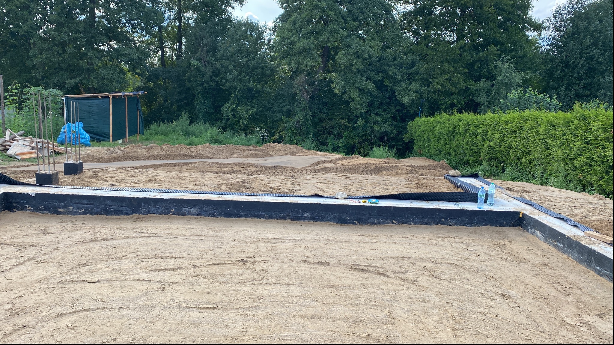 Przygotowany teren pod budowę domu z widocznymi fundamentami zabezpieczonymi czarną folią izolacyjną, dwie butelki wody stojące na fundamencie, w tle ziemia wyrównana pod wylewkę i prowizoryczna...