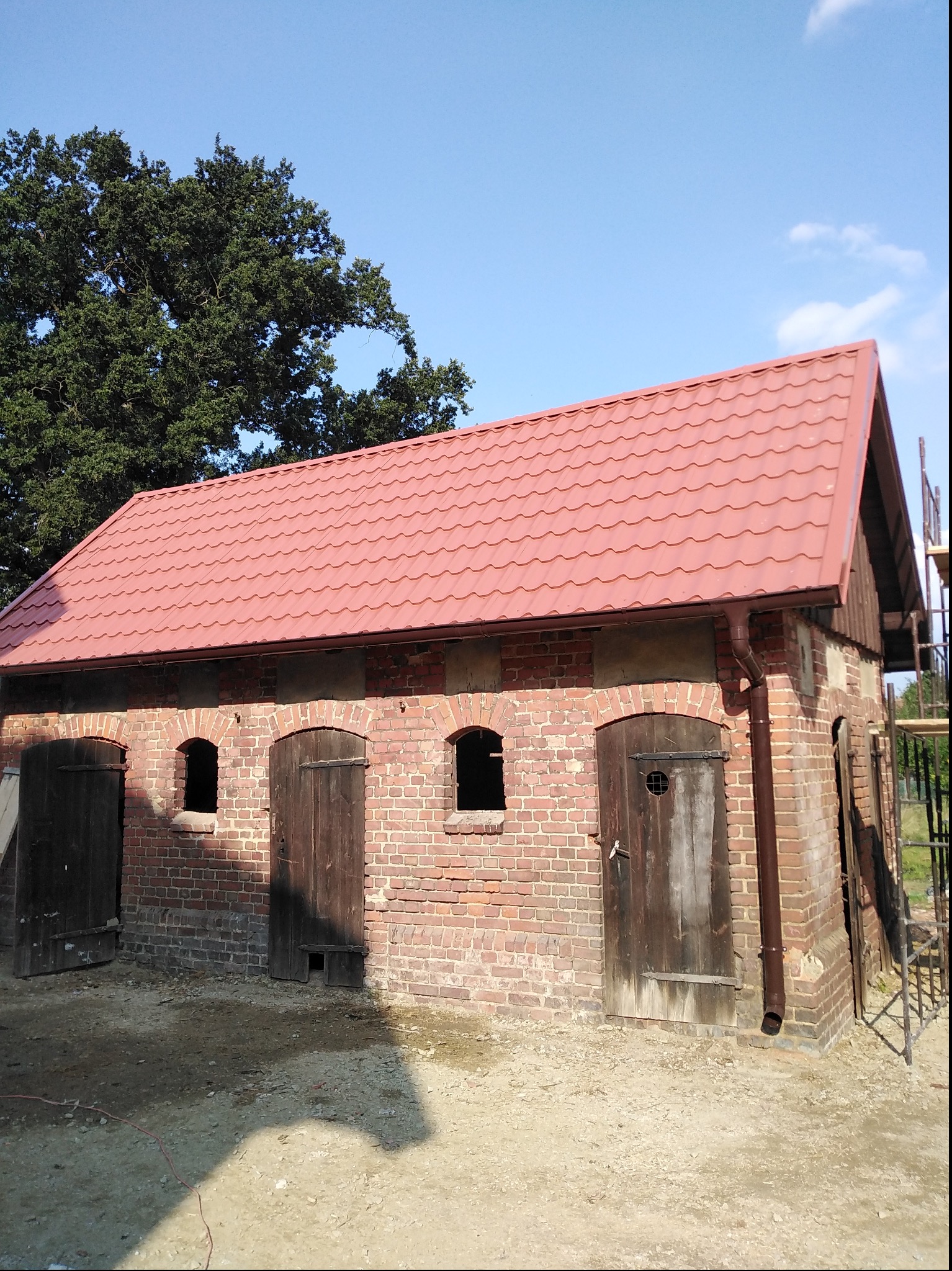 Murowany budynek gospodarczy z nowym, czerwonym dachem z blachodachówki, widoczne drewniane drzwi i okna w kształcie łuku, na zewnątrz rusztowanie.