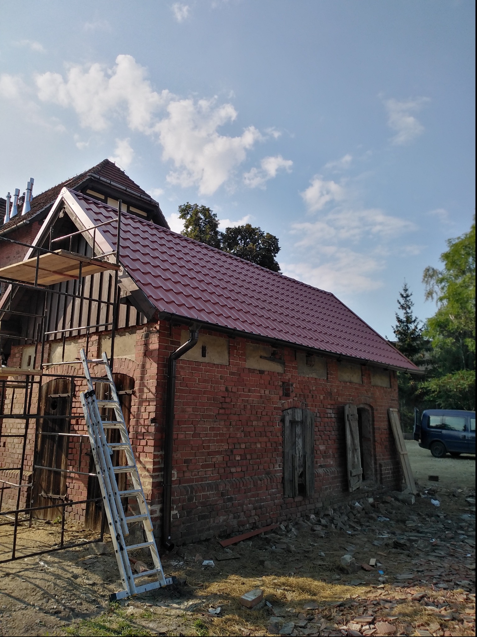 Ceglany budynek w trakcie renowacji z nowym, czerwonym dachem z dachówki; widoczne rusztowanie, drabina oparta o ścianę i gruz na ziemi.