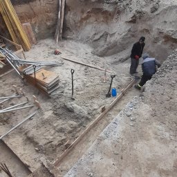 Osoba fizyczna - Dwaj pracownicy w wykopie pod fundamenty, z widocznymi deskami szalunkowymi, łopatami i stertą ziemi w tle, narzędzia leżące na ziemi.