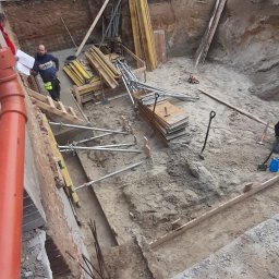 Osoba fizyczna - Głęboki wykop z drewnianym szalunkiem na obrzeżach, widoczny pracownik z planami, metalowe podpory i stosy desek, przygotowanie do wylewania fundamentów.