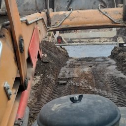 Osoba fizyczna - Widok z kabiny operatora koparki na wykop pod fundamenty z betonową wylewką, widoczny pracownik w czerwonej koszuli i drewniane szalunki.