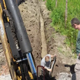Żółta koparka częściowo widoczna obok wąskiego wykopu w ziemi, gdzie mężczyzna łopatą wyrównuje dno. Drugi mężczyzna obserwuje pracę. W tle widoczny budynek i zieleń.