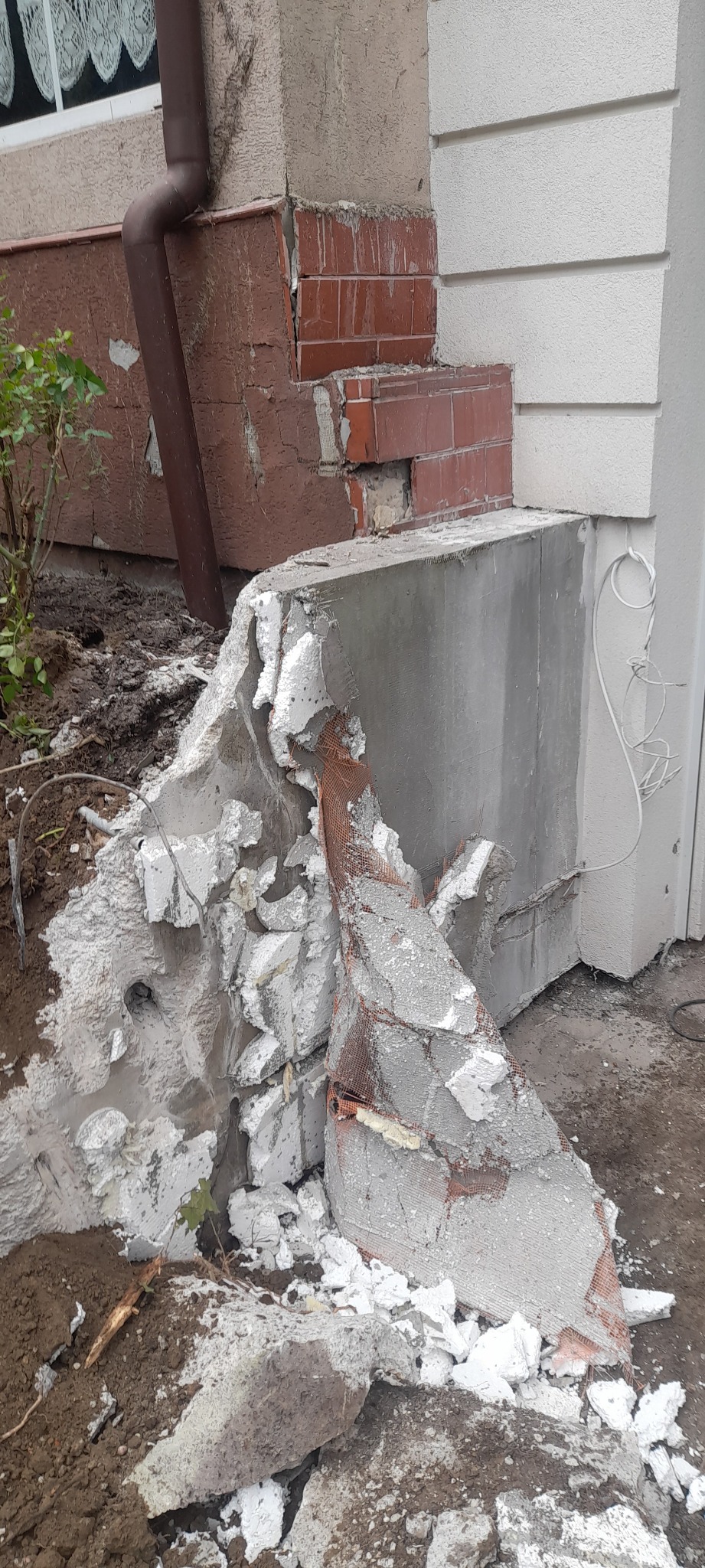 Uszkodzony narożnik budynku z odsłoniętą cegłą i fragmentami betonu oraz siatki, widoczny brązowy rynna spustowa, ziemia i fragmenty roślinności.