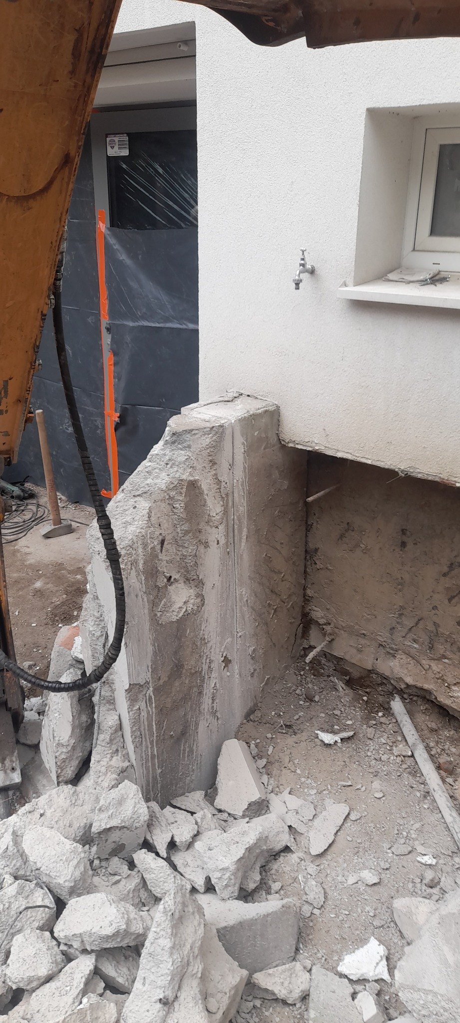 Fragment rozbiórki betonowego fundamentu przy budynku, widoczne gruzowisko i ramię koparki w tle, zabezpieczona ściana budynku folią.