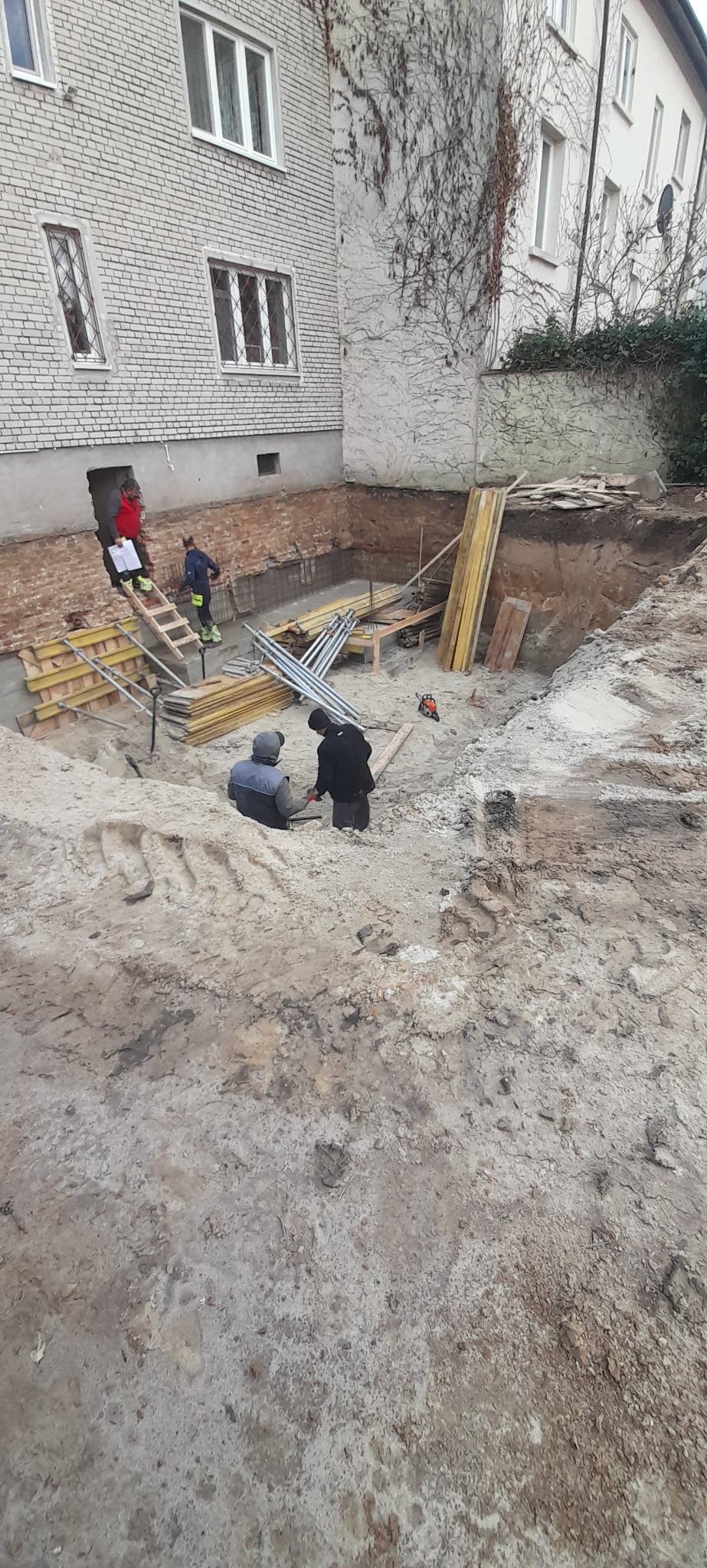 Dwóch robotników w wykopie fundamentowym przy budynku mieszkalnym, widoczne szalunki, podpory i drabina, z tyłu budynku ściana porośnięta bluszczem.