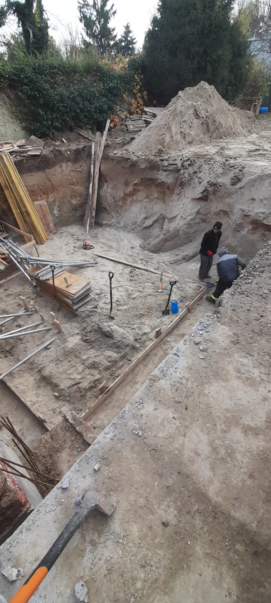 Dwaj pracownicy w wykopie pod fundamenty, z widocznymi deskami szalunkowymi, łopatami i stertą ziemi w tle, narzędzia leżące na ziemi.
