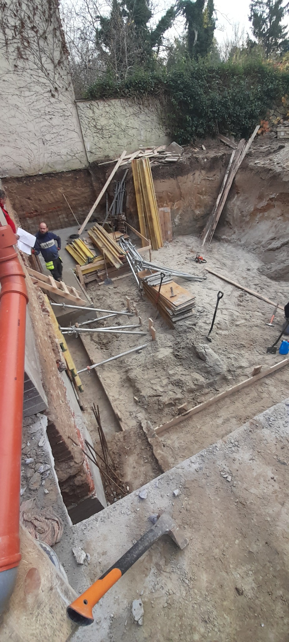 Głęboki wykop z drewnianym szalunkiem na obrzeżach, widoczny pracownik z planami, metalowe podpory i stosy desek, przygotowanie do wylewania fundamentów.