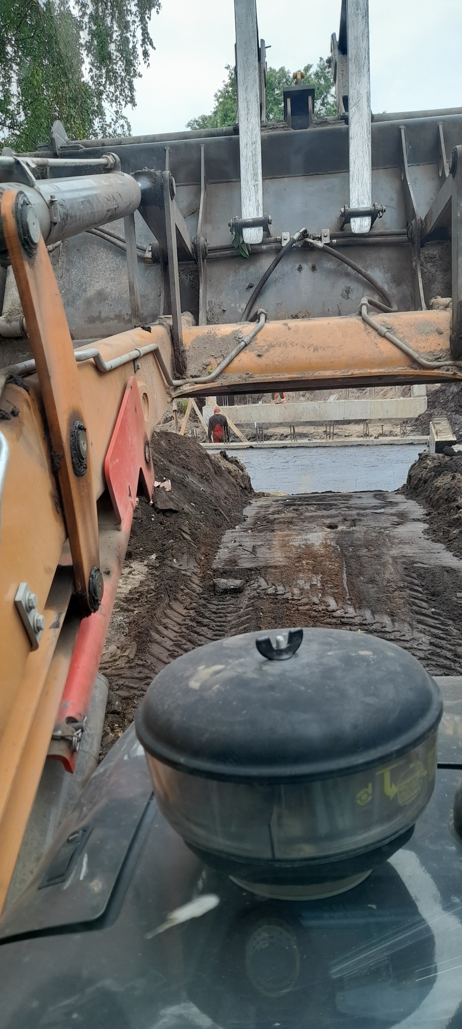 Widok z kabiny operatora koparki na wykop pod fundamenty z betonową wylewką, widoczny pracownik w czerwonej koszuli i drewniane szalunki.