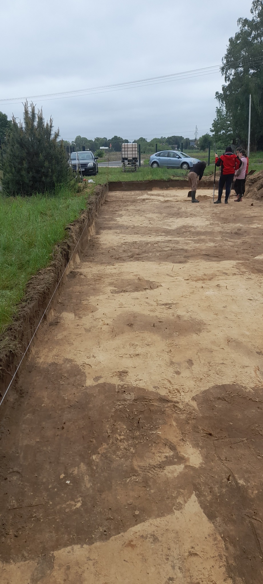 Przygotowany teren pod budowę z wykopem oznaczonym sznurkiem, w tle widoczne samochody i drzewa, pracownicy z łopatami.