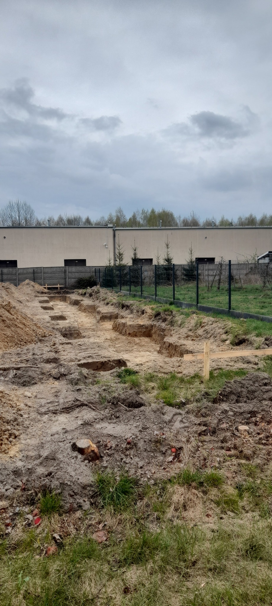 Przygotowany teren pod budowę, widoczne wykopane rowy pod fundamenty, ziemia usunięta, teren ogrodzony siatką, budynek w tle, pochmurne niebo.