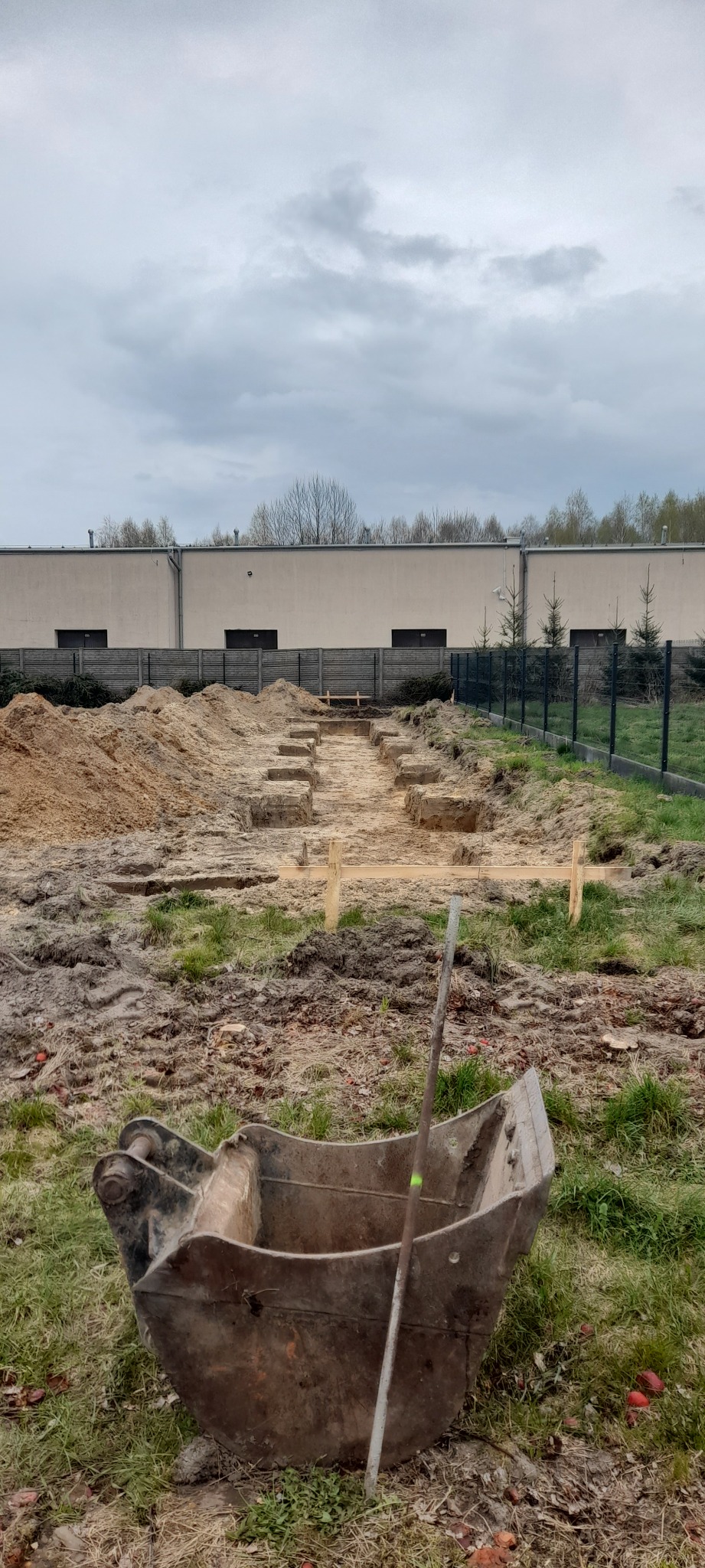 Rzędy wykopanych otworów w ziemi pod fundamenty, widoczne drewniane znaczniki, z przodu metalowa łyżka koparki, w tle budynek i ogrodzenie.