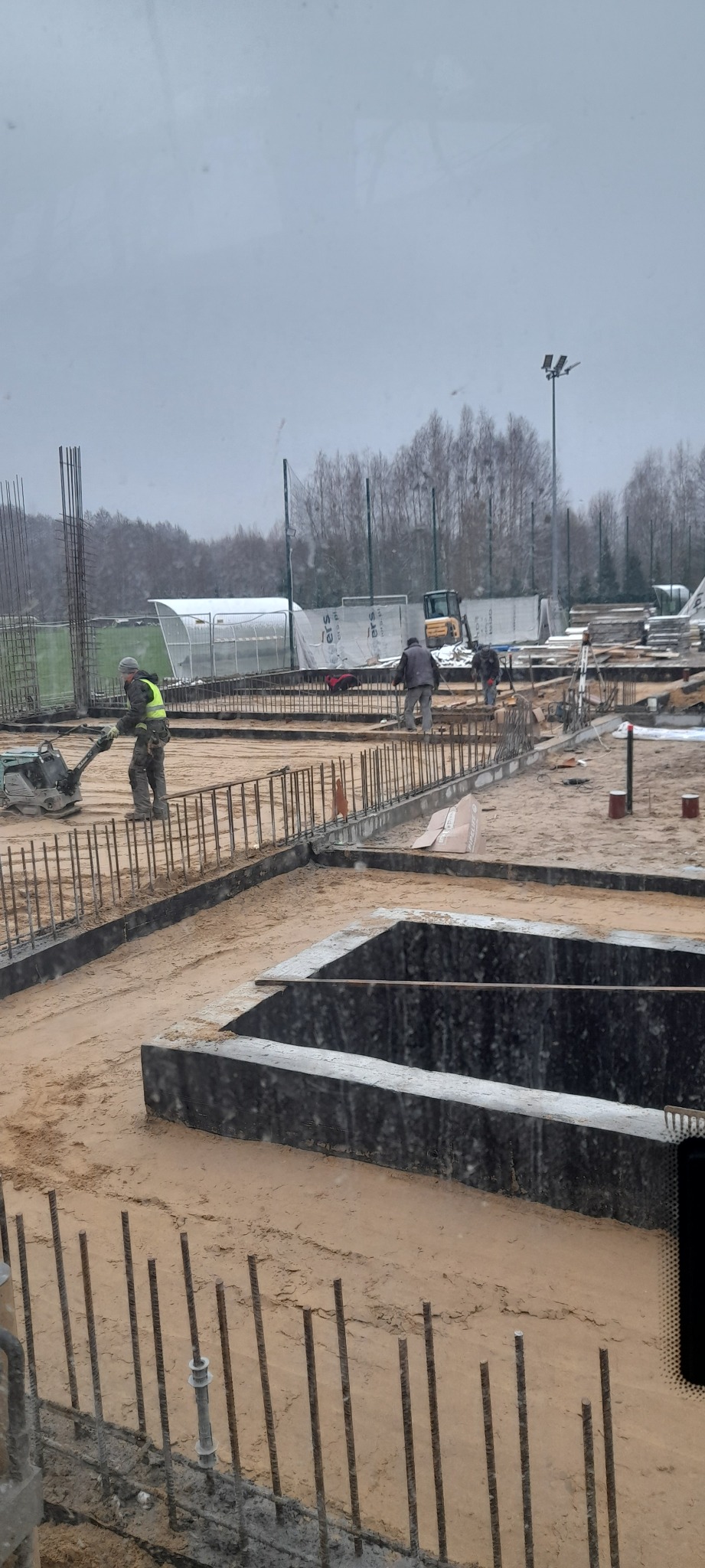 Prace ziemne w Grójcu: Robotnik zagęszcza podłoże ubijarką wibracyjną przy przygotowanych fundamentach. Widoczne zbrojenia, wykopane doły pod fundamenty i inni pracownicy na placu budowy.