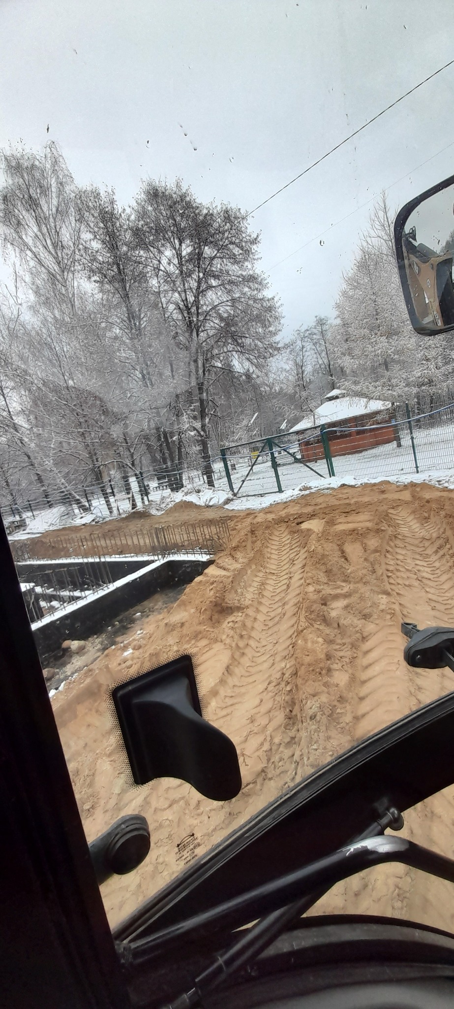 Widok z kabiny maszyny budowlanej na wykop pod fundamenty w zimowej scenerii, ślady opon na piasku, drzewa pokryte śniegiem w tle.