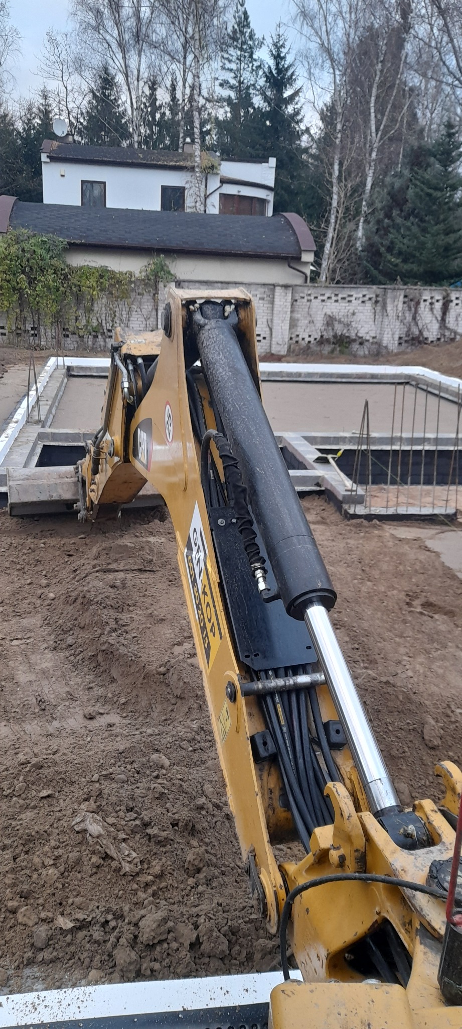 Żółta koparka CAT z ramieniem hydraulicznym na placu budowy, widoczne przygotowane szalunki fundamentów i wykopana ziemia, w tle dom jednorodzinny i drzewa.