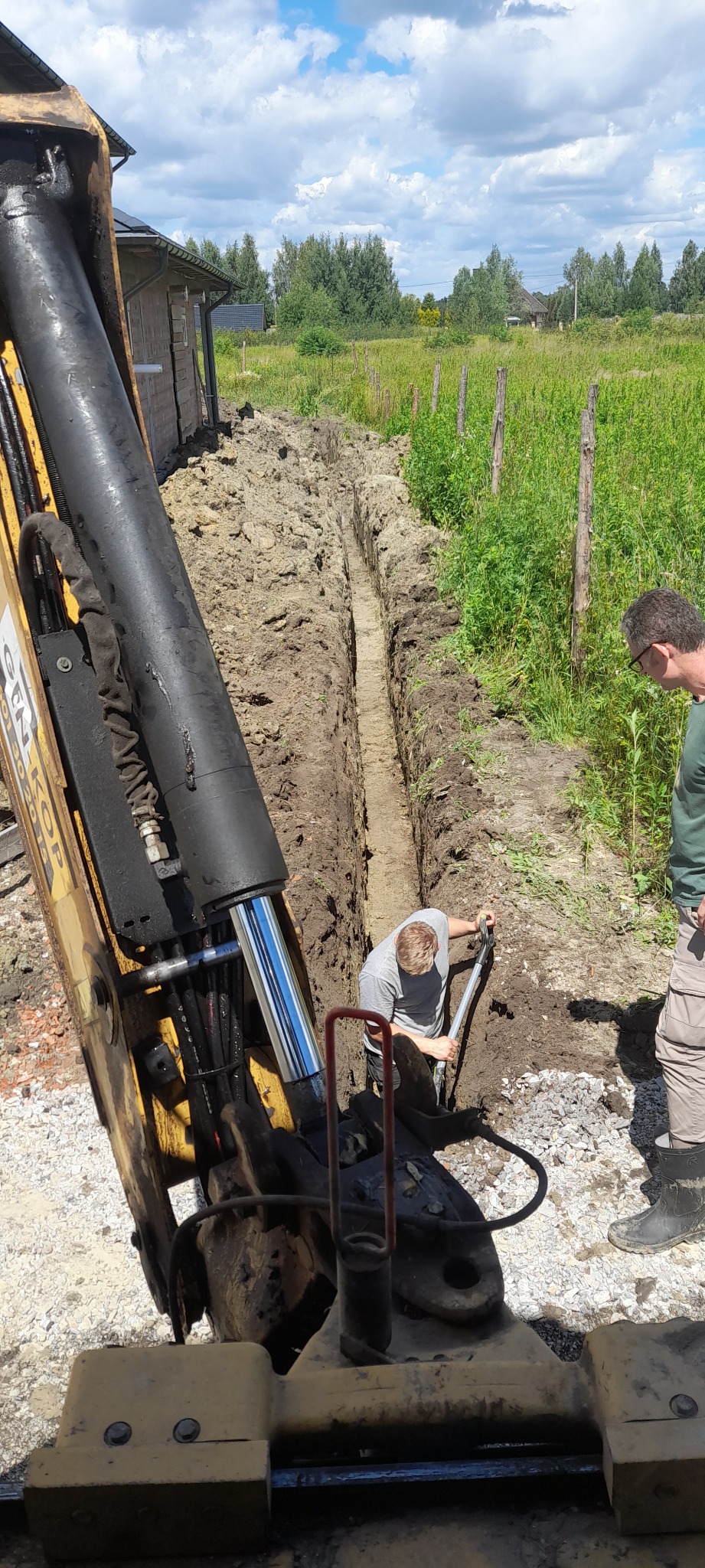 Żółta koparka częściowo widoczna obok wąskiego wykopu w ziemi, gdzie mężczyzna łopatą wyrównuje dno. Drugi mężczyzna obserwuje pracę. W tle widoczny budynek i zieleń.