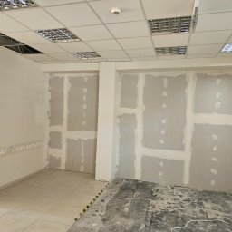 OKTAWIAN LESZCZYK - Wnętrze pomieszczenia w trakcie montażu płyt kartonowo-gipsowych, widoczne łączenia płyt zaszpachlowane, sufit podwieszany z oświetleniem rastrowym, fragment podłogi z płytek i fragment z surowego...