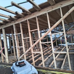 OKTAWIAN LESZCZYK - Konstrukcja drewniana domu w trakcie budowy, widoczne elementy szkieletu dachu i ścian, narzędzia budowlane w tle.