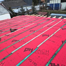 OKTAWIAN LESZCZYK - Montaż łat na czerwonej membranie dachowej z nadrukowanym logo, widoczne narzędzia i fragment sylwetki pracownika w pomarańczowych butach, w tle budynki i drzewa.