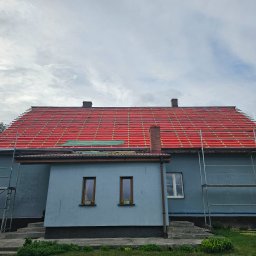 OKTAWIAN LESZCZYK - Dom z częściowo zdjętym starym dachem i przygotowanym podkładem z czerwonej membrany dachowej. Widoczne rusztowania i dwa kominy. Etap prac przygotowawczych.