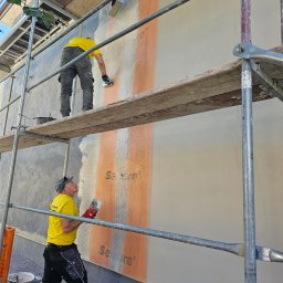 OKTAWIAN LESZCZYK - Dwóch pracowników na rusztowaniu aplikuje zaprawę na ocieploną ścianę budynku. Widoczne narzędzia i materiały budowlane. Etap prac wykończeniowych elewacji.