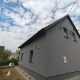 Szary dom jednorodzinny po renowacji elewacji, widok z boku, z czarnym dachem i elementami instalacji na ścianie, na tle błękitnego nieba z chmurami.