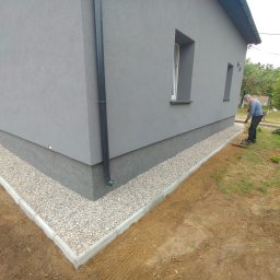 OKTAWIAN LESZCZYK - Szara elewacja domu z wykończonym cokołem i kamiennym obramowaniem. Mężczyzna grabi ziemię obok budynku. Widok z niskiego kąta.