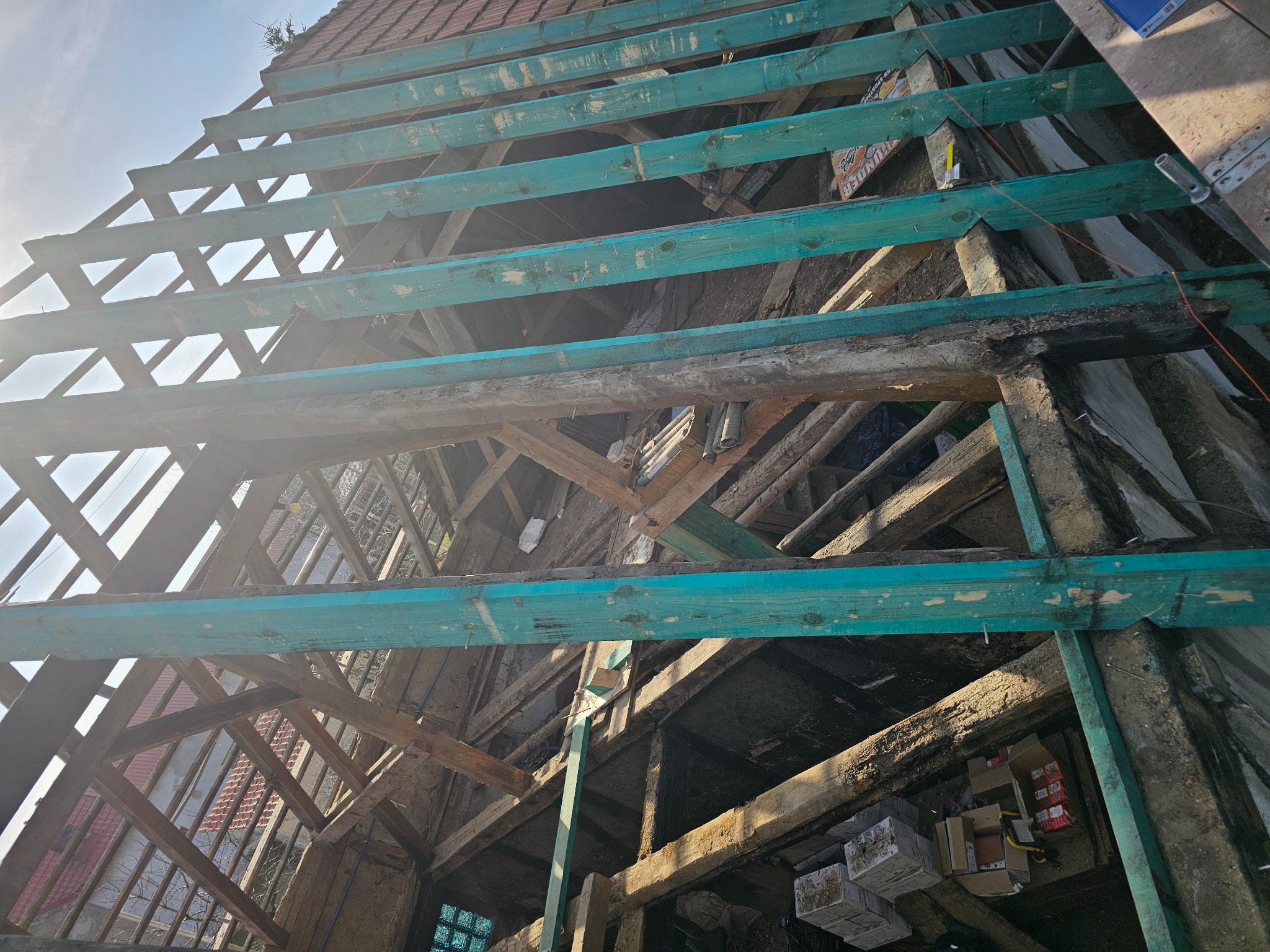 Drewniana konstrukcja dachu w trakcie remontu, zabezpieczona turkusowymi belkami, widok od wewnątrz, fragment starego dachu z dachówką w tle.
