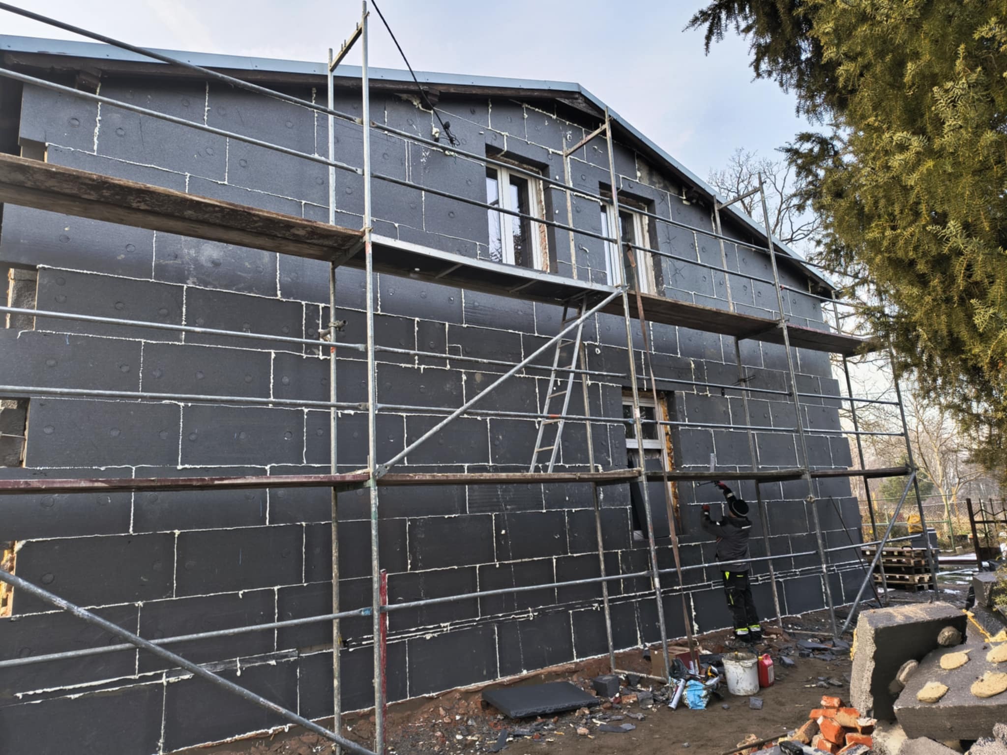 Dom w trakcie ocieplania, widoczne czarne płyty izolacyjne na elewacji, rusztowanie i pracownik przy oknie, przygotowujący elewację do dalszych prac wykończeniowych.