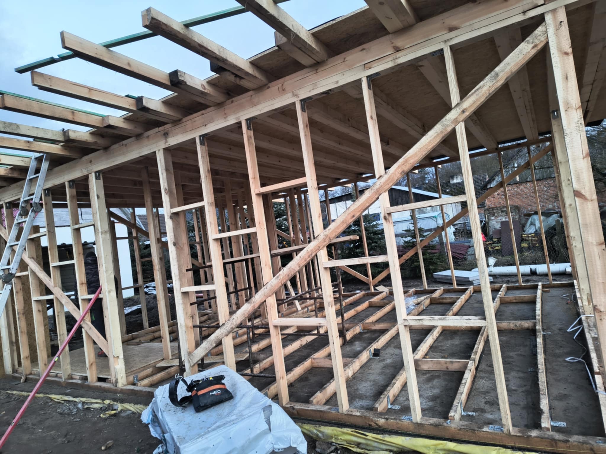 Konstrukcja drewniana domu w trakcie budowy, widoczne elementy szkieletu dachu i ścian, narzędzia budowlane w tle.
