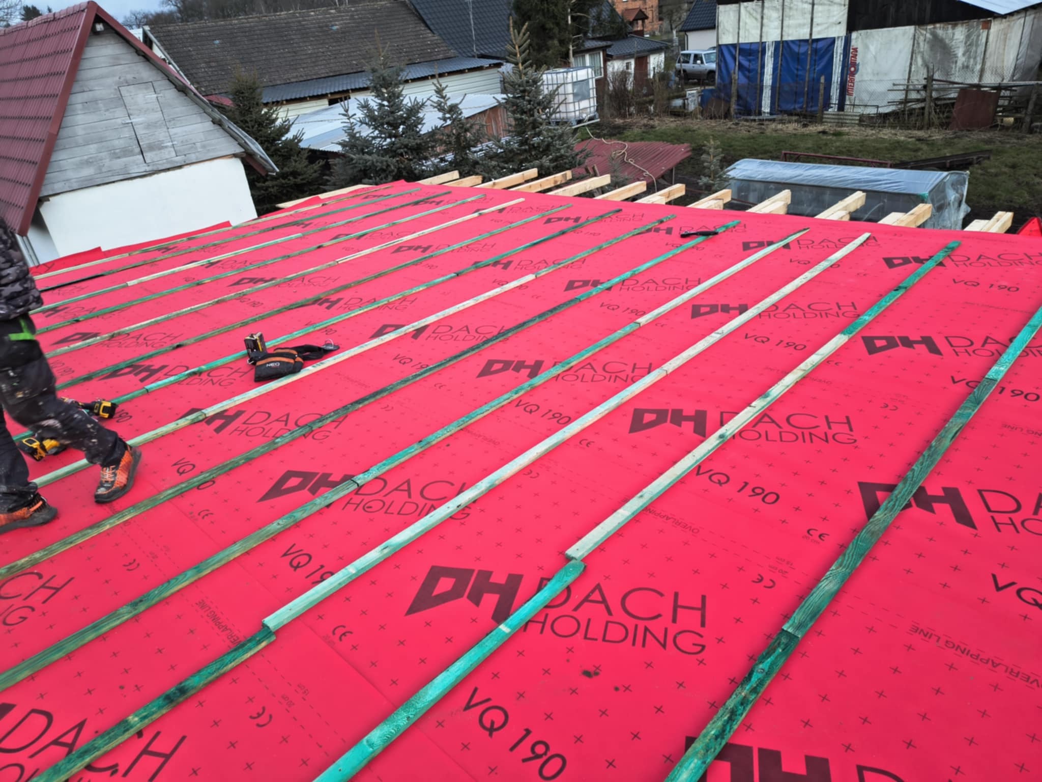 Montaż łat na czerwonej membranie dachowej z nadrukowanym logo, widoczne narzędzia i fragment sylwetki pracownika w pomarańczowych butach, w tle budynki i drzewa.