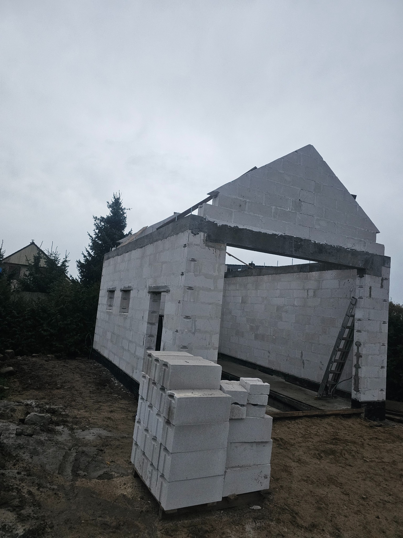 Wznoszony budynek z bloczków betonowych, widoczne ściany, otwory okienne i fragment dachu szczytowego, na pierwszym planie stos bloczków, w tle drzewa i pochmurne niebo.