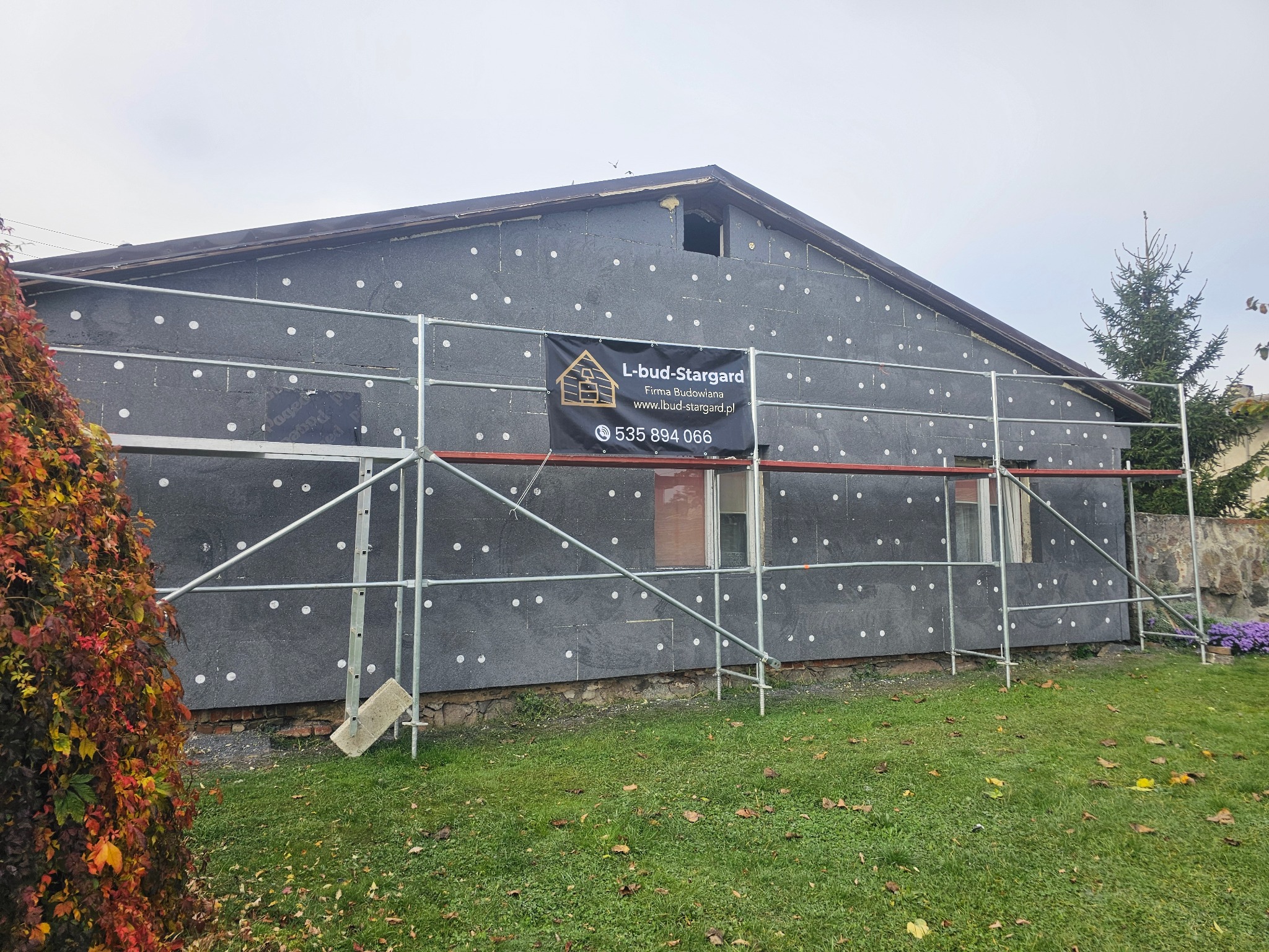 Dom w trakcie prac związanych z izolacją termiczną elewacji, widoczne rusztowanie, szara płyta izolacyjna z białymi kołkami mocującymi oraz baner reklamowy firmy budowlanej.